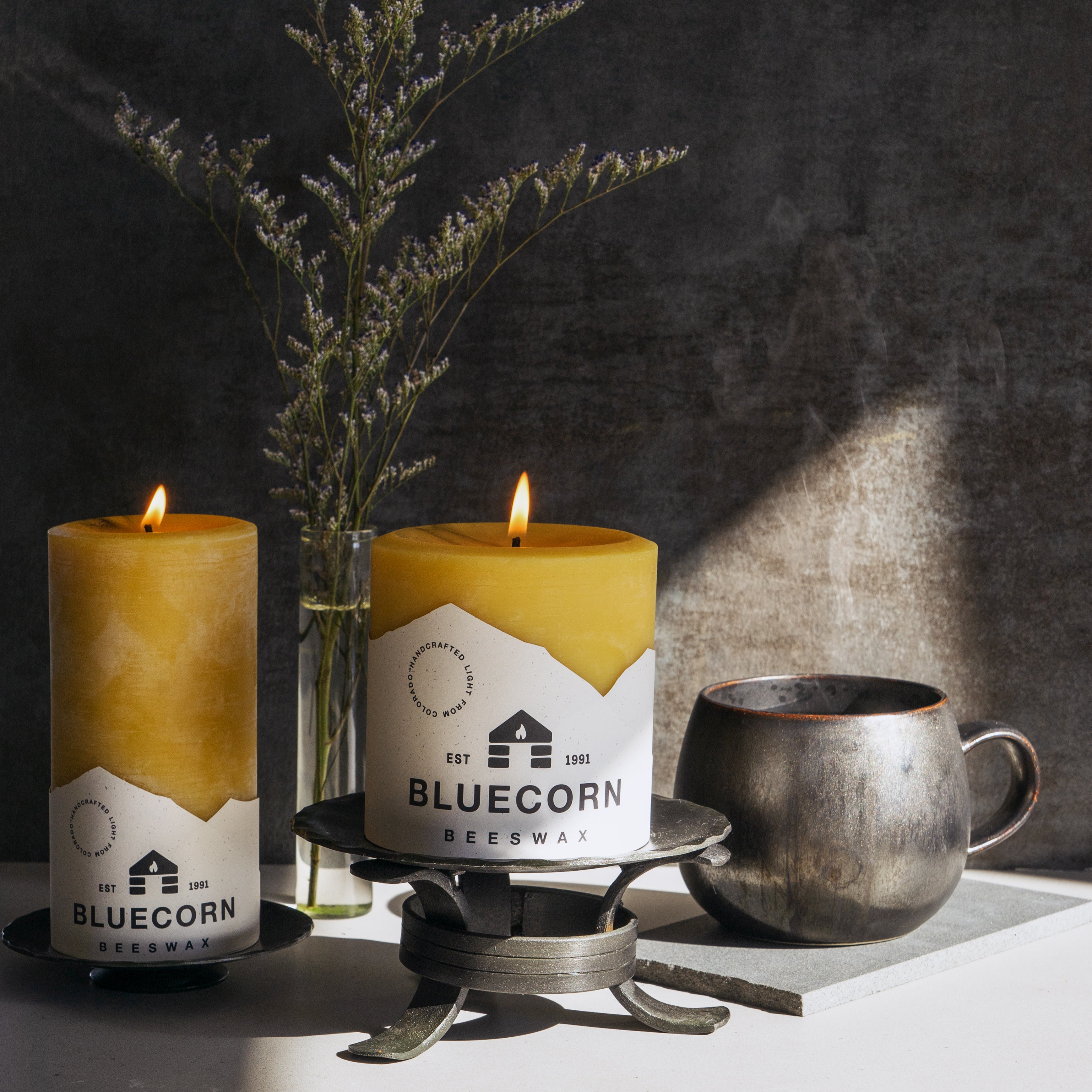 Two lit candles with 'Bluecorn' branding, a vase with dried plants, and a metal mug on a dark background.