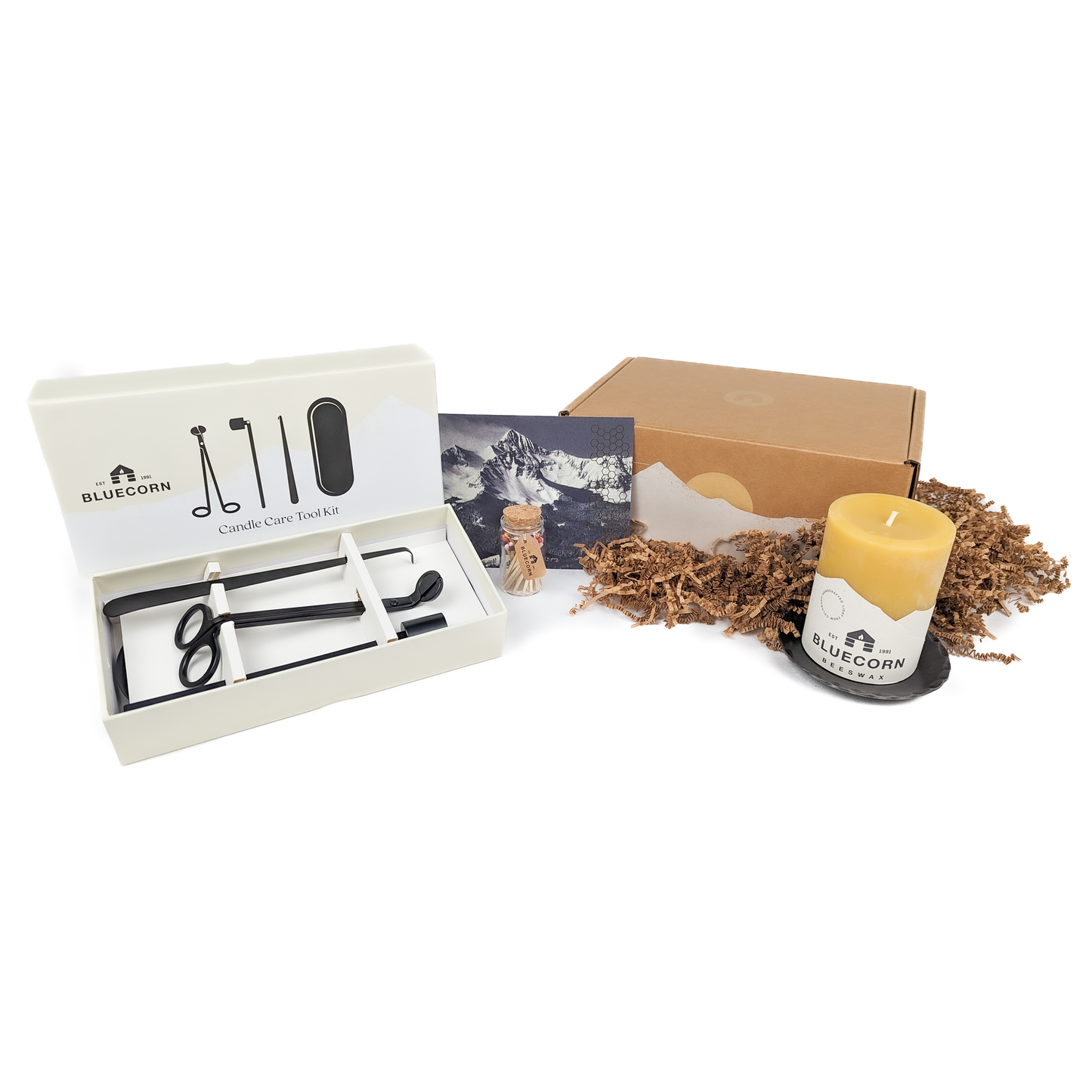 A Pure Beeswax Pillar Gift Kit with a gold-foiled gift box, including a 3" x 4" pure beeswax pillar candle, a 4.75" hand-forged pillar base, a candle care toolkit, a refillable apothecary jar of rainbow matches, and a gift card with a handwritten message.