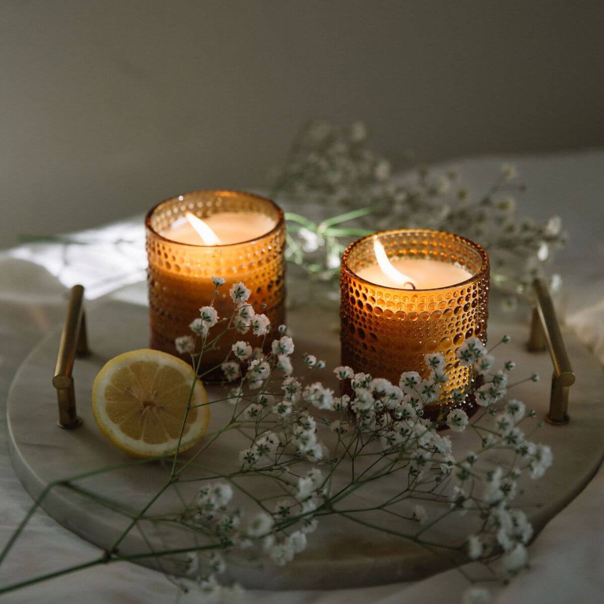 Two lit candles in textured glass holders on a marble tray with flowers and a lemon.