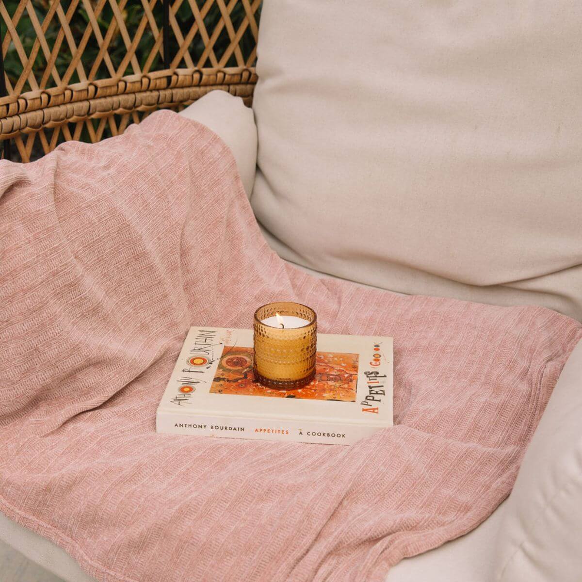 Cozy scene with a lit scented candle on a stack of books, draped with a soft pink blanket.
