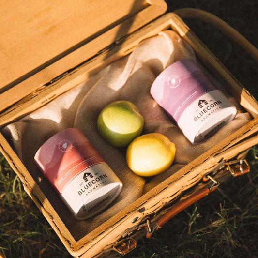 Scented candle jars in a picnic basket with fresh lemons and green apples outdoors.
