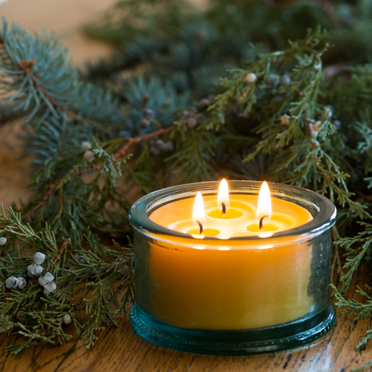 Beeswax scented candle with three wicks, surrounded by evergreen branches on a wooden table.
