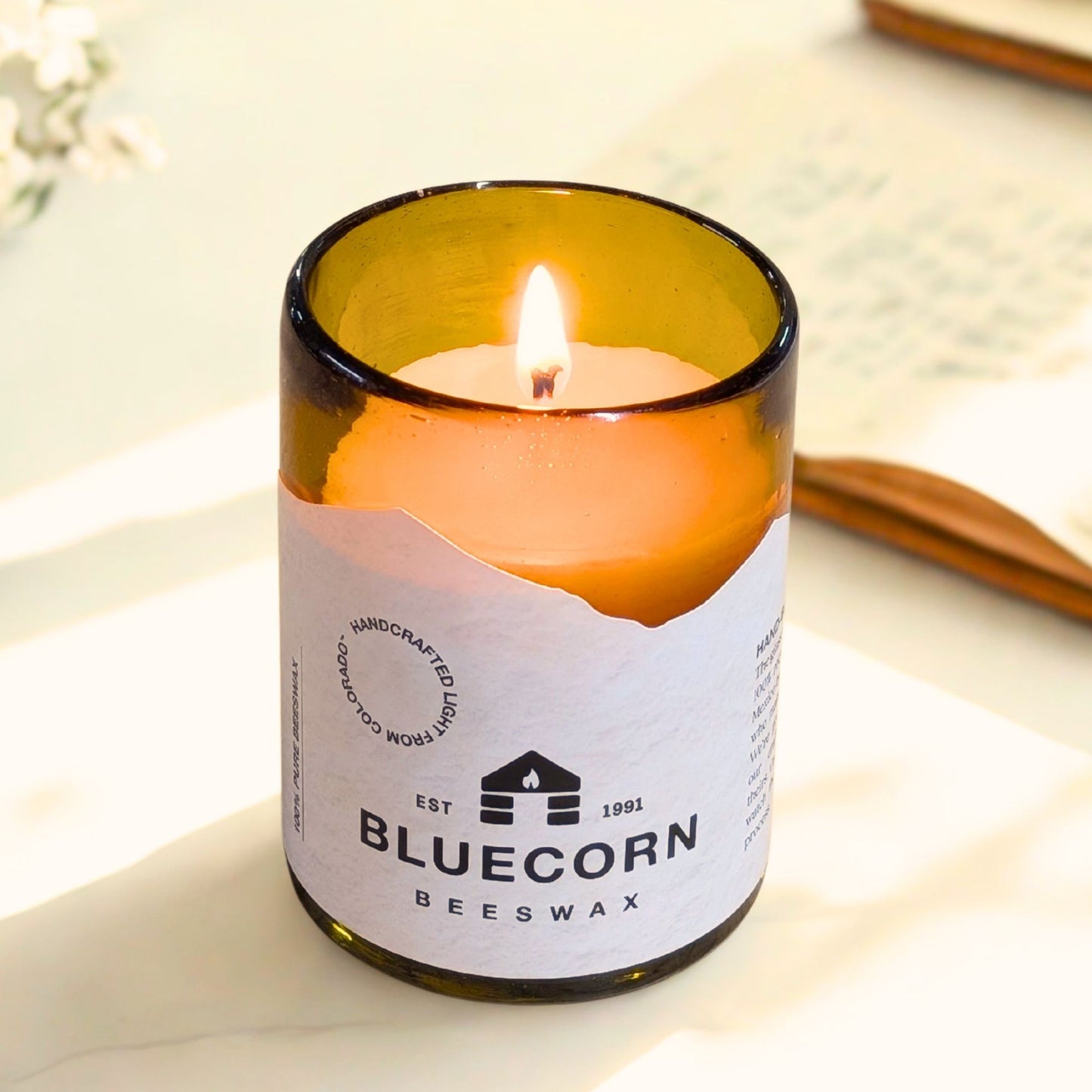 Candle in a glass bottle labeled 'Bluecorn Beeswax' on a light surface.