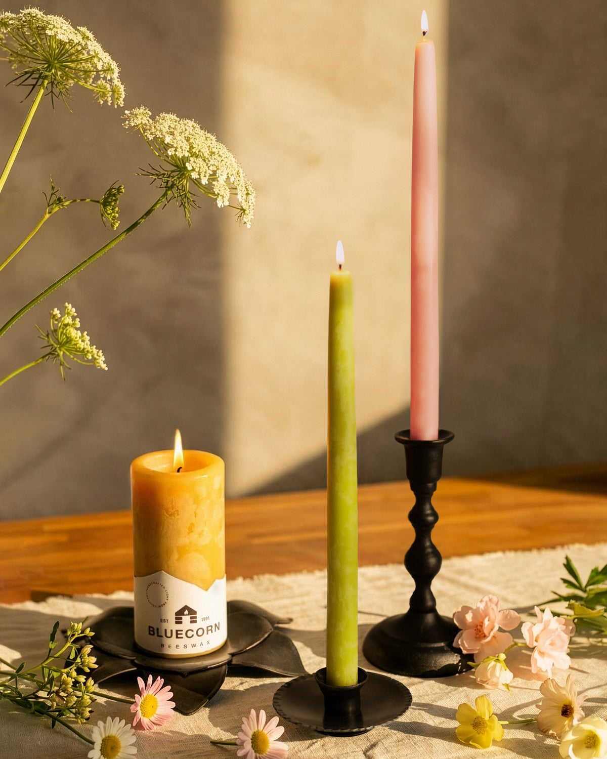 Beeswax taper candles in elegant candlesticks with a honey-scented candle, surrounded by fresh flowers.