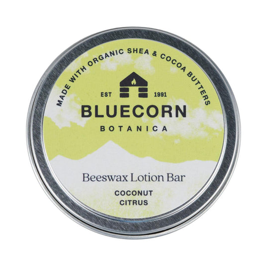 Bluecorn Botanica Beeswax Lotion Bar in Coconut Citrus. Made with Organic Shea Butter, Organic Cocoa Butter, Avocado Oil, Apricot Oil, Cappings Beeswax, Essential Oils. Using the warmth of your hands, these solids bars will turn into lotions. Can be used all over the body and come in a convenient tin. Features green Bluecorn Botanica Label on aluminum tin with opened lid showing lotion bar. Lotion bar is cream in color and features Bluecorn cabin.