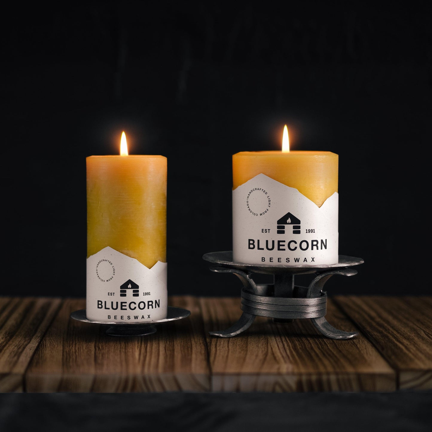 Two burning beeswax pillar candles atop forged pillar holders in front of a black backdrop.