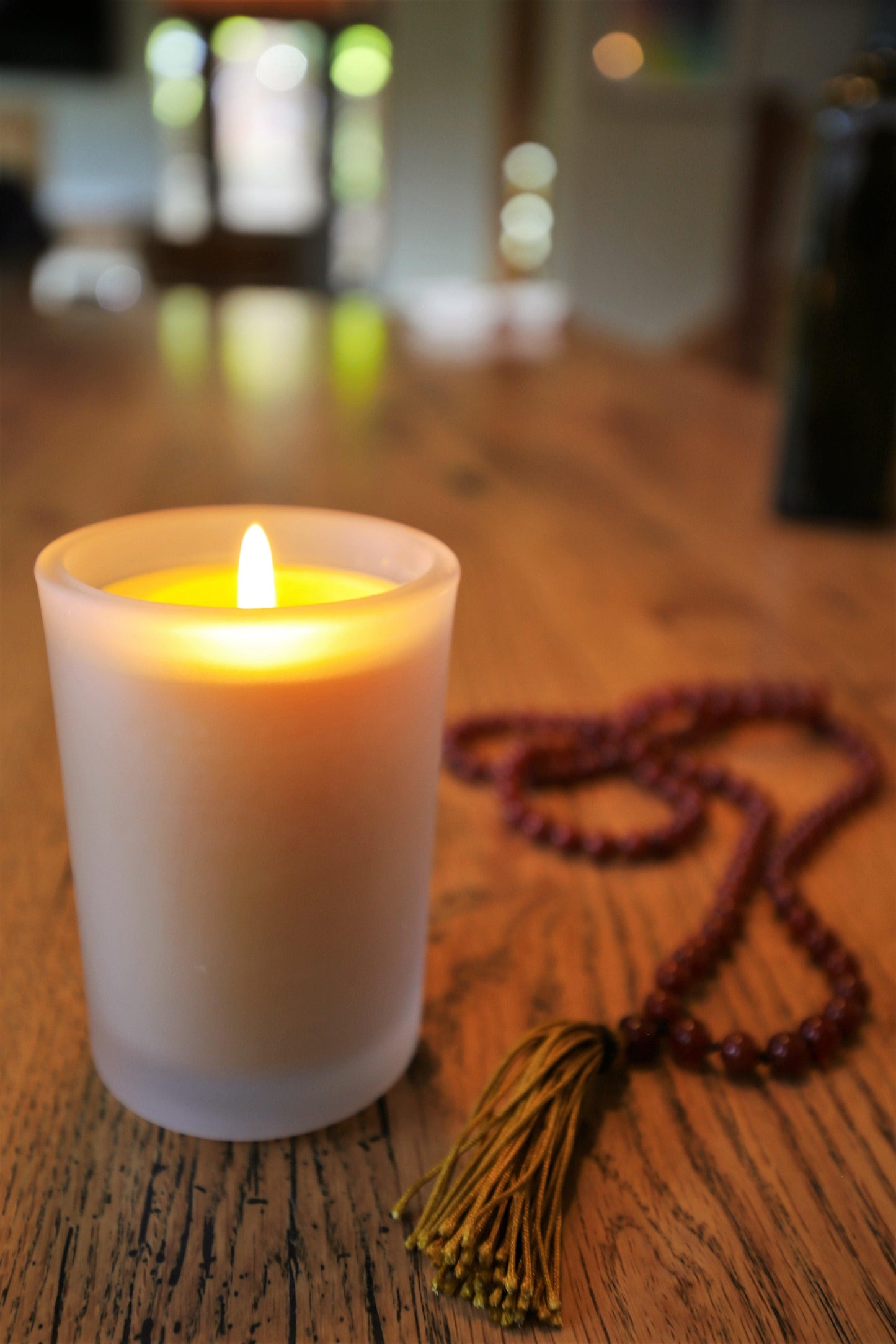 Lit Bluecorn Beeswax 8.5oz frosted candle. Candle is on wooden table and red mala next to it. Wax is golden in color and in shows through only slightly in the frosted glass.