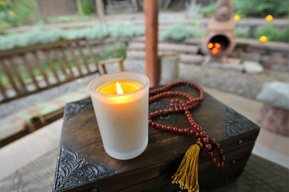 Lit Bluecorn Beeswax 8.5oz frosted candle outdoors. Candle is on wooden chest and red mala next to it, a bench and garden is slightly shown in the background. Wax is golden in color and in shows through only slightly in the frosted glass.