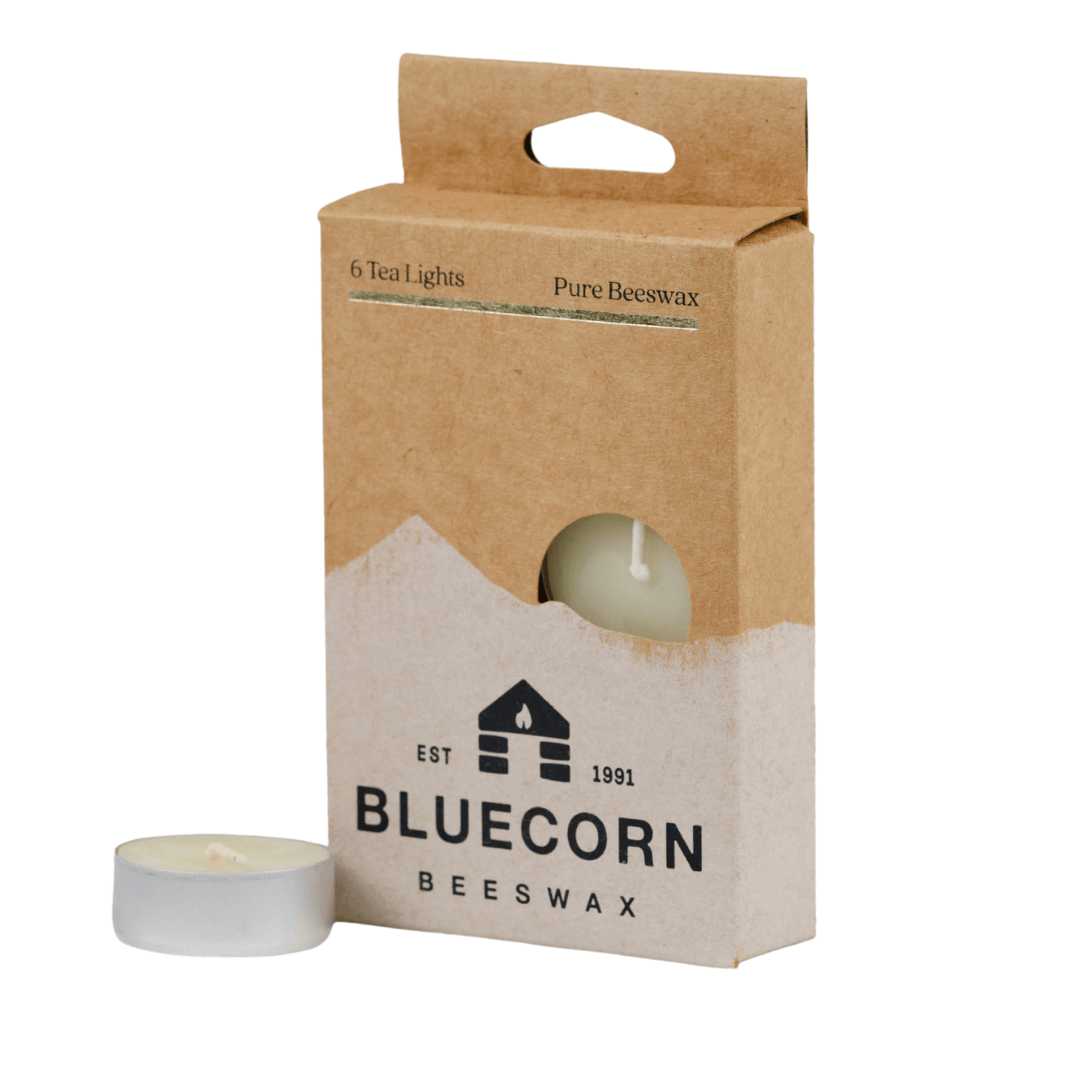 Bluecorn Beeswax 100% Pure Beeswax 6 Pack Ivory Tea Light in Metal cups. Picture features one Tea Light outside of Bluecorn branded Tea Light Box with a white background. Box features Bluecorn Beeswax name and logo, with an outline of Mt. Wilson in white printed on a 100% recycled kraft paper box. Tea lights will burn for about 5 hours each.