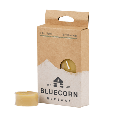 Bluecorn Beeswax 100% Pure Beeswax 6 Pack Raw Tea Light in Clear cups. Picture features one Tea Light outside of Bluecorn branded Tea Light Box with a white background. Box features Bluecorn Beeswax name and logo, with an outline of Mt. Wilson in white printed on a 100% recycled kraft paper box. Tea lights will burn for about 5 hours each.