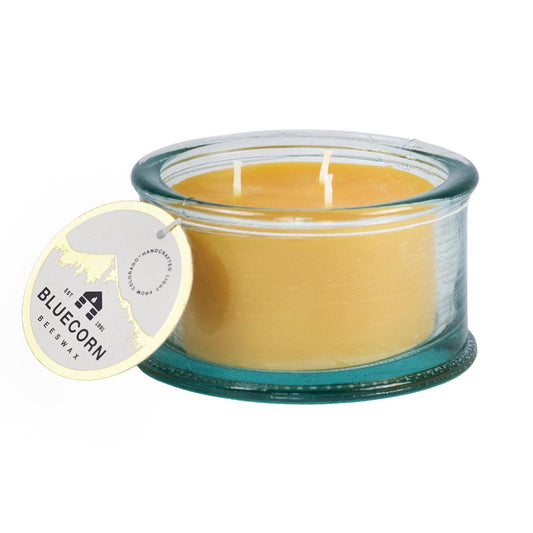Image displays Bluecorn Beeswax 3-Wick candle in 100% recycled Spanish glass. The wax is golden in color and the glass is a watery aqua color. Candle has hang tag with Bluecorn name and logo and an outline of the mountains in gold foil.