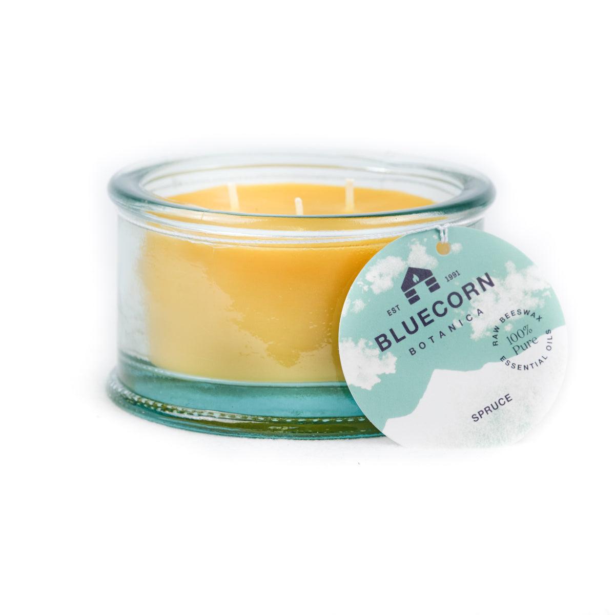 Bluecorn Botanica 100% Pure Beeswax Spruce 10oz 3-Wick Glass Candle. Wax is golden in color and glass is clear colored, with a white back drop. Features green Bluecorn Botanica hang tag printed on 100% recyled paper with Bluecorn name and logo. Burn Time: 18 hours.  Made with 100% Pure Beeswax, 100% Pure Essentail Oils and a 100% pure cotton wick, no lead. Candles are paraffin free, clean burning and non-toxic. 