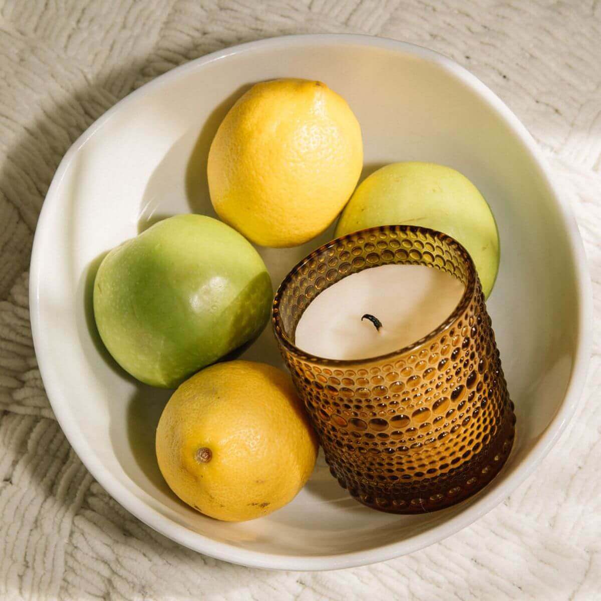 Scented candle in a textured amber jar surrounded by green and yellow fruits in a white bowl.