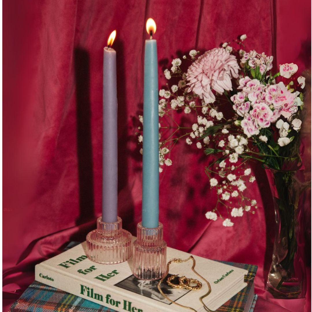 Two lit beeswax taper candles in glass holders on books with a bouquet of flowers against a red curtain.
