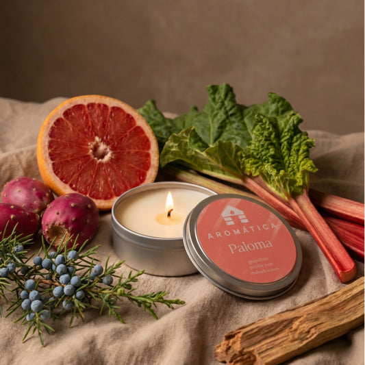 A lit silver tin candle labeled Aromática Paloma sits on draped linen fabric, surrounded by natural ingredients including a sliced grapefruit, rhubarb stalks, blueberries, rosemary, and wood bark. The product is styled with its lid beside the open candle.