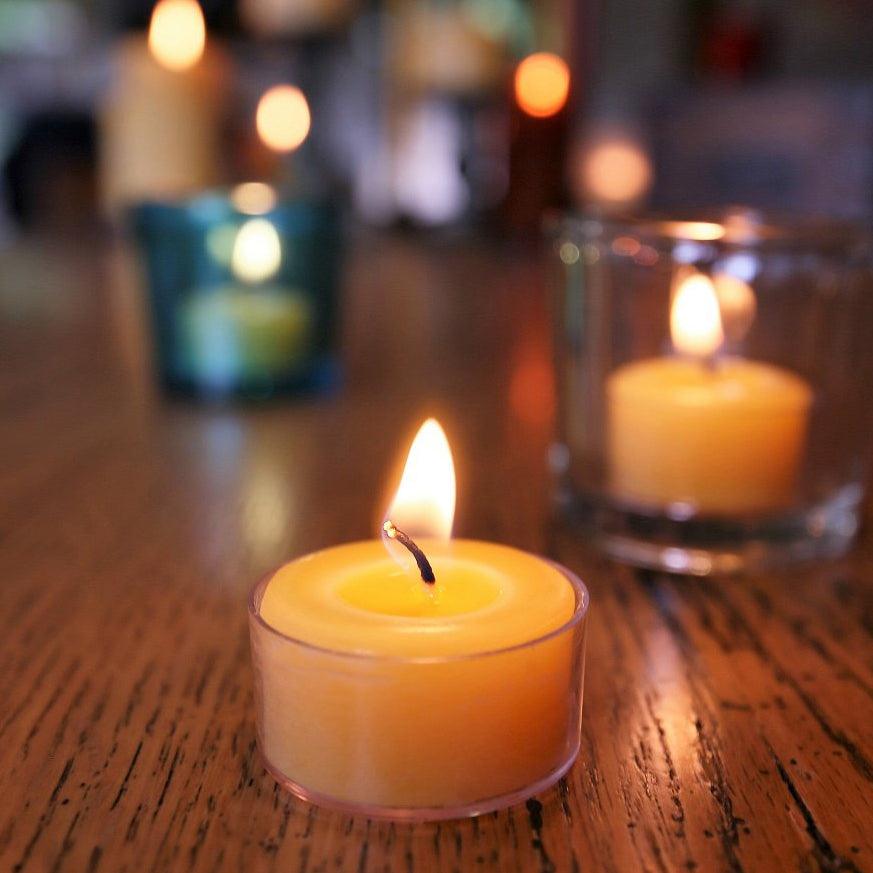 A beeswax tea light in a poly-carbonate cup is in focus, with various types of beeswax candles burning in the background.