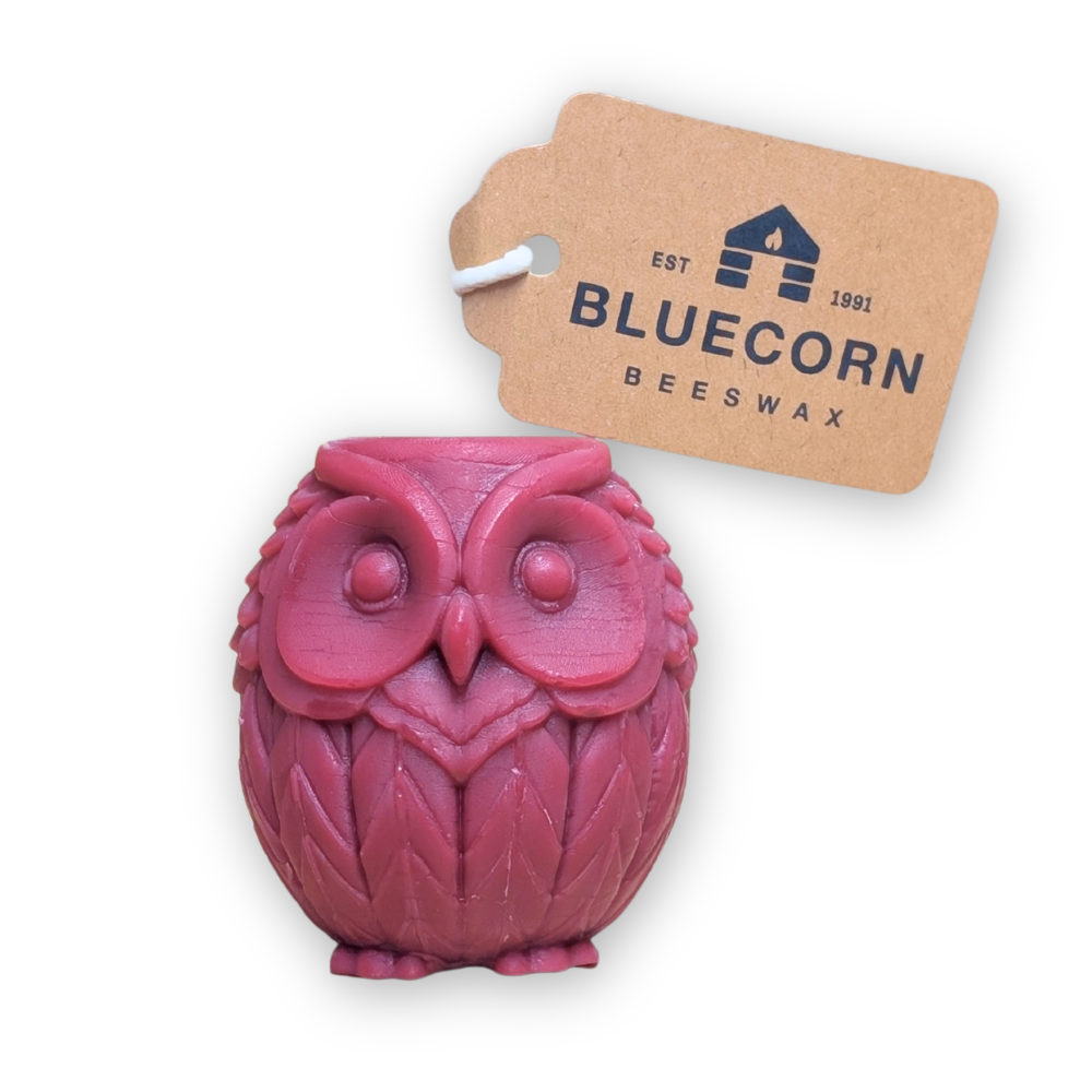 A burgundy owl-shaped beeswax candle with a textured feather pattern, shown next to its brown kraft paper tag printed with the brand 'BLUECORN BEESWAX' 
