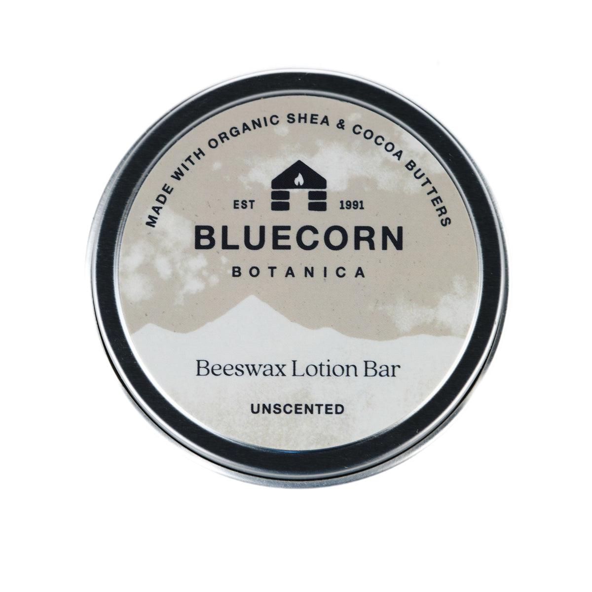Bluecorn Botanica Beeswax Lotion Bar in Unscented. Made with Organic Shea Butter, Organic Cocoa Butter, Avocado Oil, Apricot Oil, Cappings Beeswax, Essential Oils. Using the warmth of your hands, these solids bars will turn into lotions. Can be used all over the body and come in a convenient tin. Features grey Bluecorn Botanica Label on aluminum tin with opened lid showing lotion bar. Lotion bar is cream in color and features Bluecorn cabin.