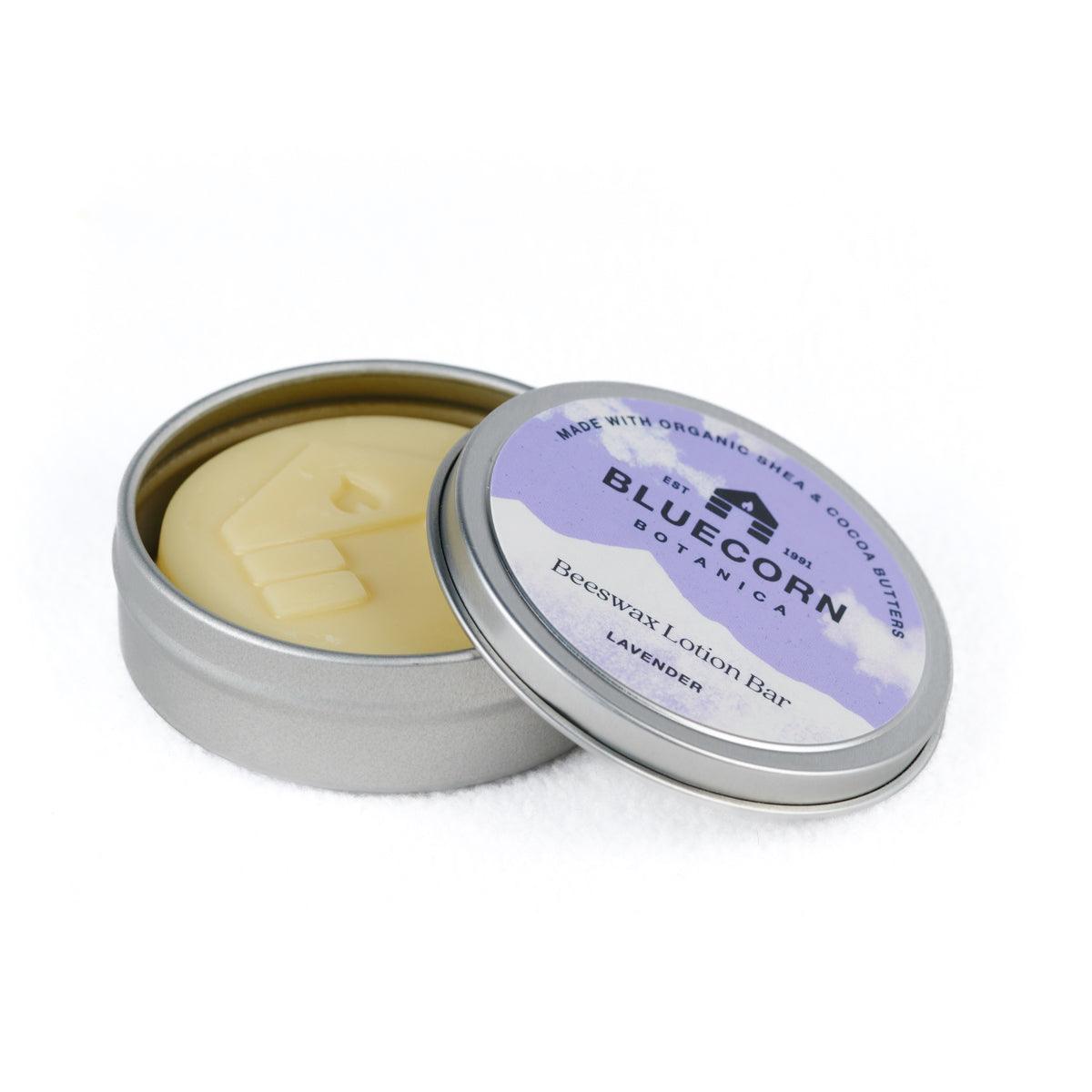 Bluecorn Botanica Beeswax Lotion Bar in Lavender. Made with Organic Shea Butter, Organic Cocoa Butter, Avocado Oil, Apricot Oil, Cappings Beeswax, Essential Oils. Using the warmth of your hands, these solids bars will turn into lotions. Can be used all over the body and come in a convenient tin. Features Purple Bluecorn Botanica Label on aluminum tin with opened lid showing lotion bar. Lotion bar is cream in color and features Bluecorn cabin. 