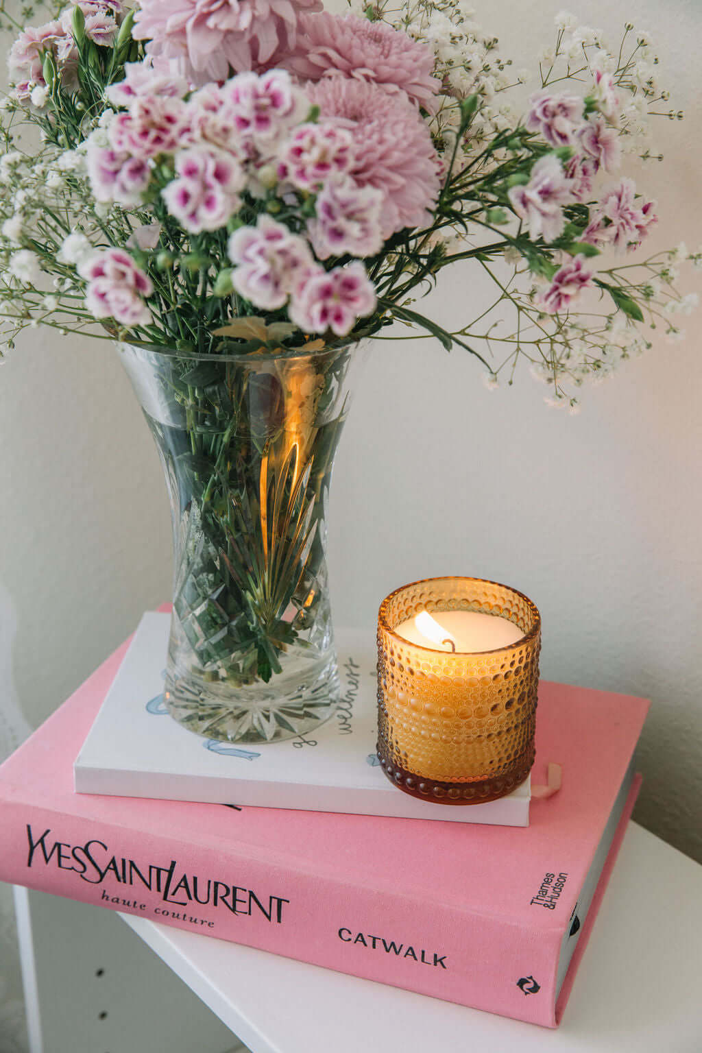 Pink flowers in a vase on a pink book with a lit scented candle from bluecorn