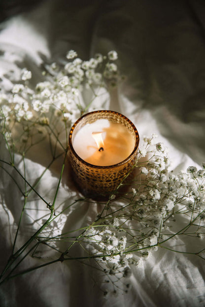 Candle in a textured holder surrounded by small white flowers on a soft surface