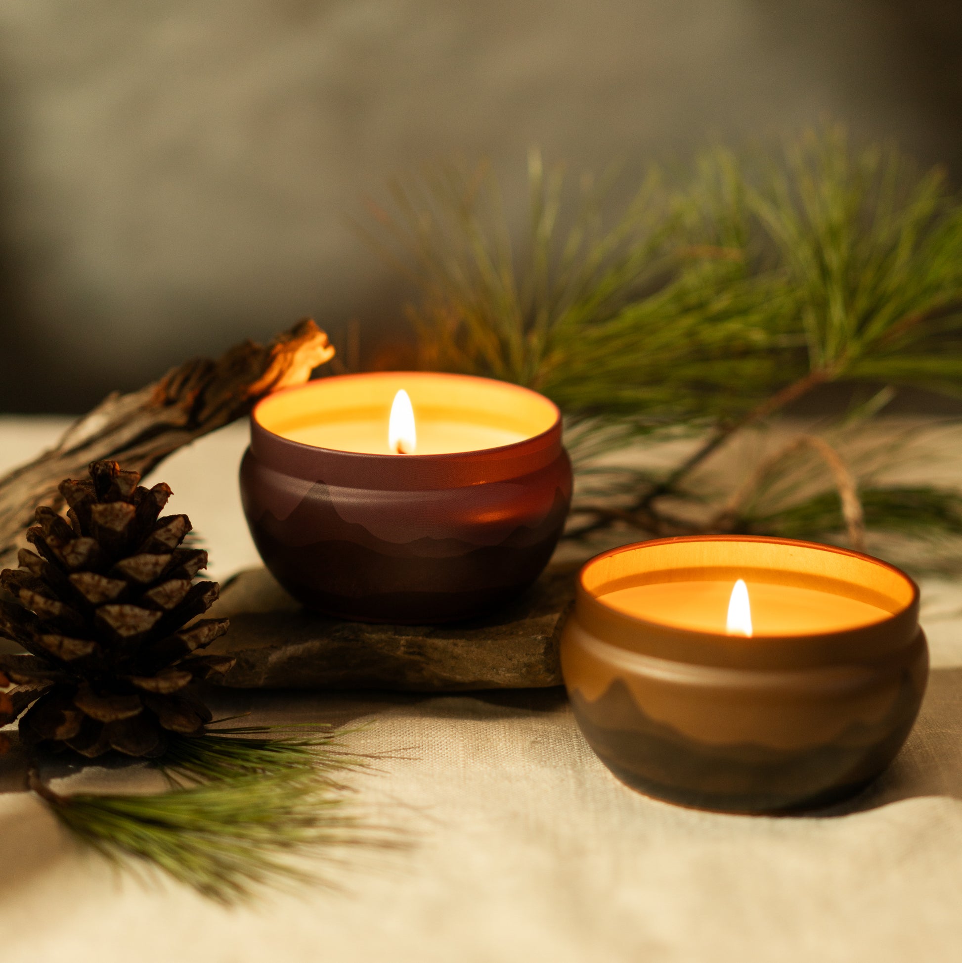 two lit candle tins bluecorn aromatica with mountain range on side burgundy and orange 