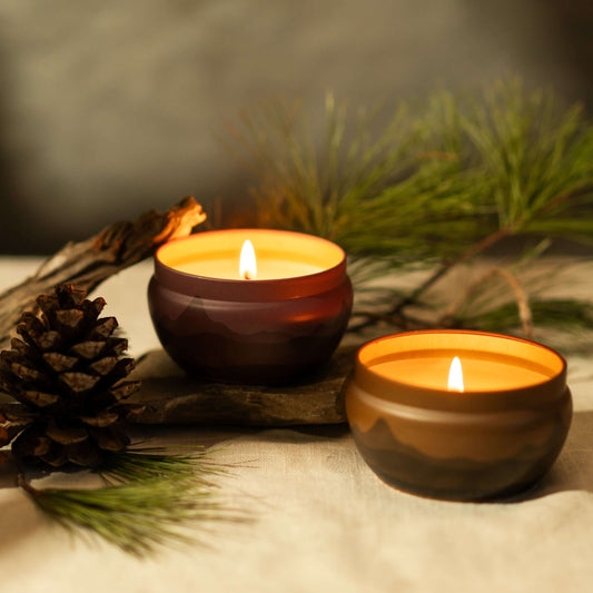 two lit scented candle tins bluecorn aromatica with mountain range on side burgundy and orange