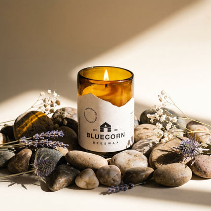 Bluecorn Beeswax candle in an amber glass container on a stone surface with lavender flowers.