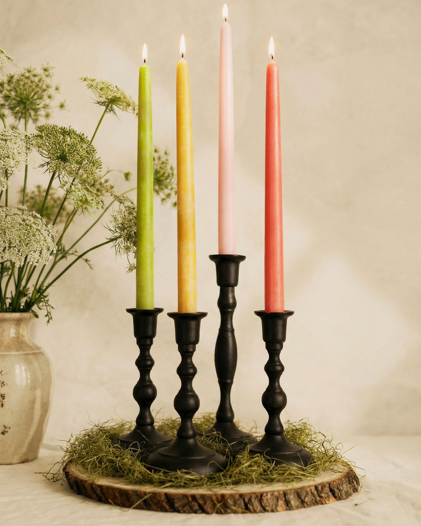 Four lit candles in black candlesticks on a wooden base with a vase of flowers in the background.