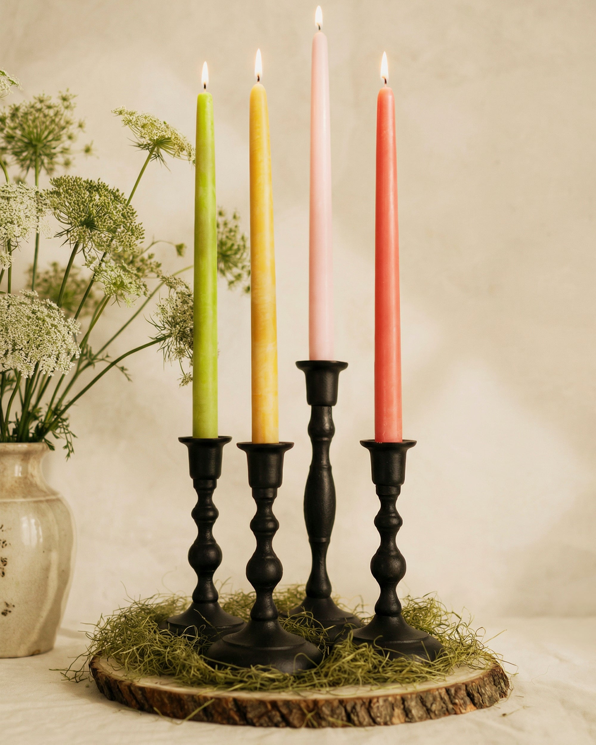 Four lit candles in black candlesticks on a wooden base with a vase of flowers in the background.