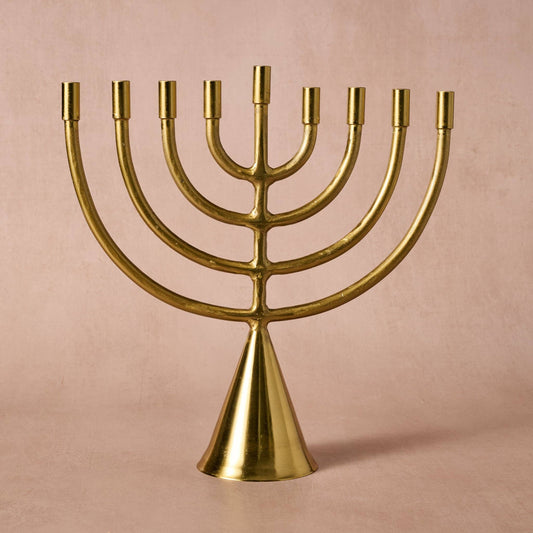 A polished brass nine-branched candelabra (menorah) with a conical base, set against a solid, warm beige-pink background.