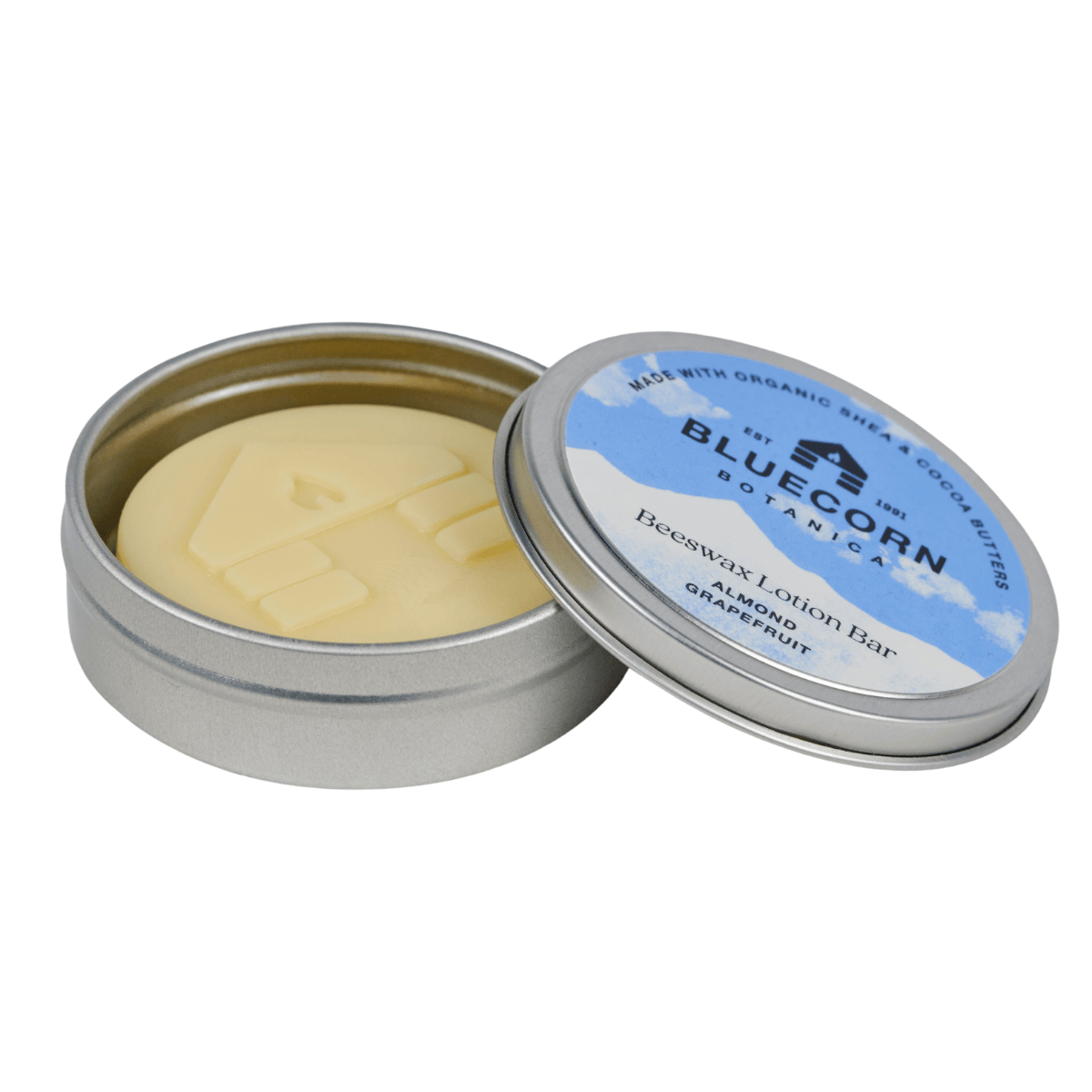 Bluecorn Botanica Beeswax Lotion Bar in Almond Grapefruit. Made with Organic Shea Butter, Organic Cocoa Butter, Avocado Oil, Apricot Oil, Cappings Beeswax, Essential Oils. Using the warmth of your hands, these solids bars will turn into lotions. Can be used all over the body and come in a convenient tin. Features blue Bluecorn Botanica Label on aluminum tin with opened lid showing lotion bar. Lotion bar is cream in color and features Bluecorn cabin.