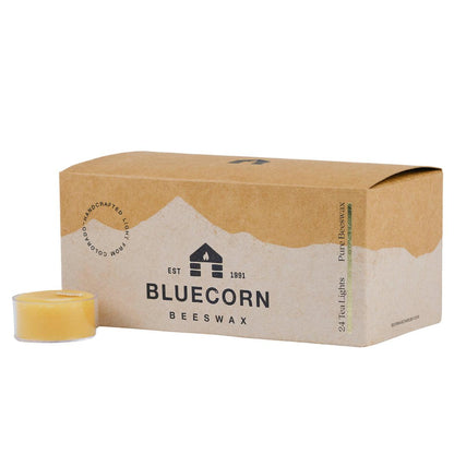 Bluecorn Beeswax 100% Pure Beeswax 24 Pack Raw Tea Light in Clear cups. Picture features one Tea Light outside of Bluecorn branded Tea Light Box with a white background. Box features Bluecorn Beeswax name and logo, with an outline of Mt. Wilson in white printed on a 100% recycled kraft paper box. Tea lights will burn for about 5 hours each.