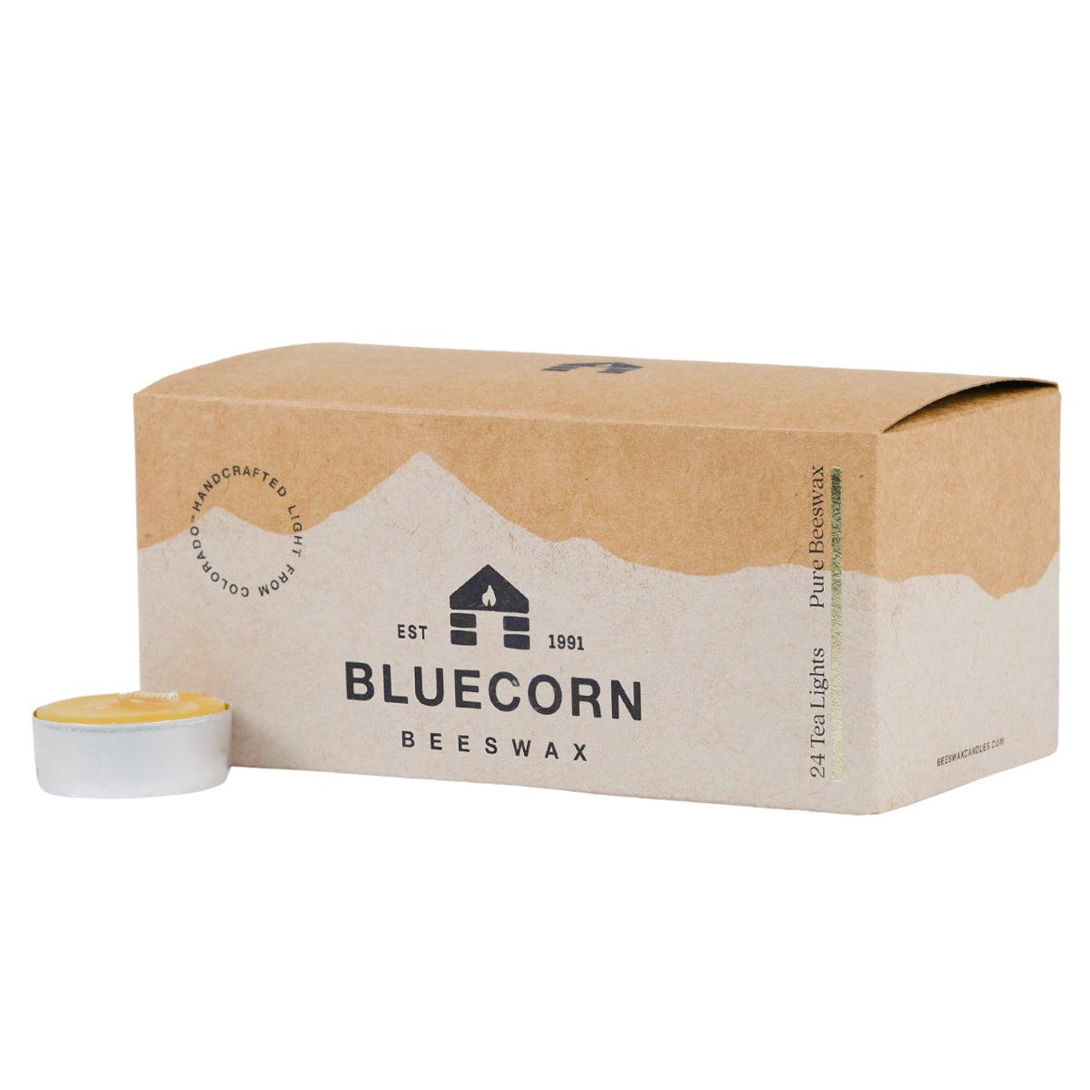Bluecorn Beeswax 100% Pure Beeswax 24 Pack Raw Tea Light in Metal cups. Picture features one Tea Light outside of Bluecorn branded Tea Light Box with a white background. Box features Bluecorn Beeswax name and logo, with an outline of Mt. Wilson in white printed on a 100% recycled kraft paper box. Tea lights will burn for about 5 hours each.