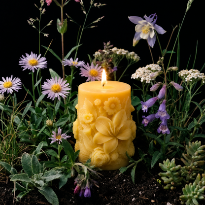 A yellow pillar candle with embossed floral details and a small flame, surrounded by purple wildflowers, green foliage, and dark soil against a black background.