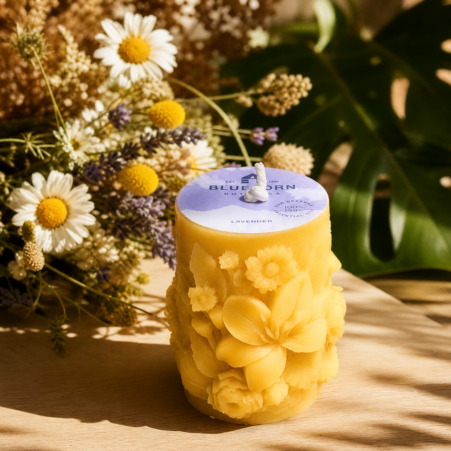 A yellow decorative candle with embossed flowers and a lavender label, styled on a wooden surface with dried flowers and green leaves in warm sunlight. pure beeswax and essential oil candle from bluecorn candles