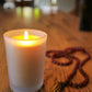 Lit Bluecorn Beeswax 8.5oz frosted candle. Candle is on wooden table and red mala next to it. Wax is golden in color and in shows through only slightly in the frosted glass.