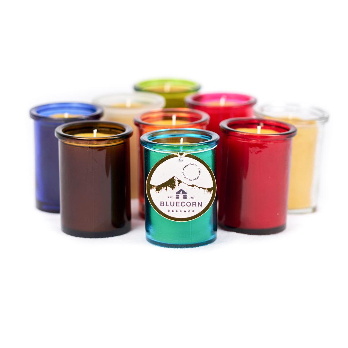 Group of Bluecorn Beeswax 6oz glass candles with Bluecorn hand tag featuring name, logo, and outline of mountains in gold foil. Colors include; Aqua, Red, Clear Fuchsia, Orange, Lime, Frosted, Cobalt, and Dark Amber. Wax is golden color. 