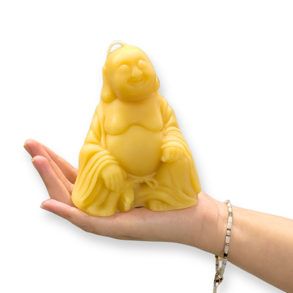 A raw pure beeswax molded buddha candle resting on a person's open hand, shown against a plain white background.