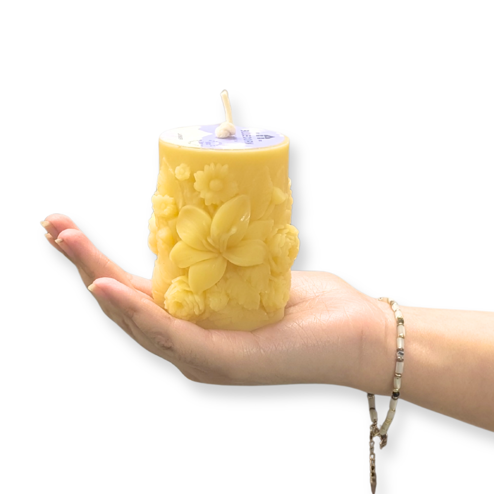 A yellow pillar candle with embossed floral details, held in the palm of a hand against a plain white background.