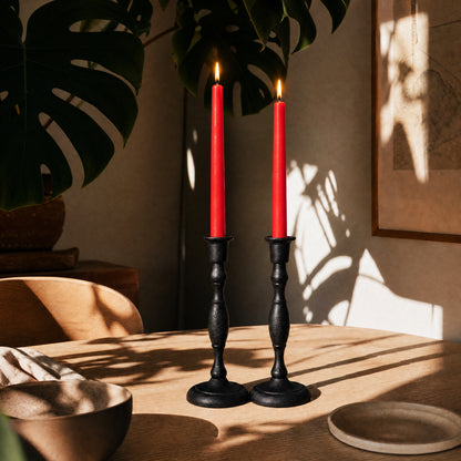 Two red candles in black candlesticks on a wooden table with a plant and wall art in the background.