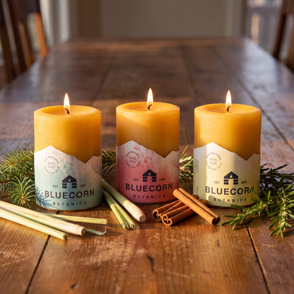 Three lit Bluecorn Botanica beeswax pillar candles in warm yellow wax are arranged on a rustic wooden table with pine branches, cinnamon sticks, and lemongrass. The labels show the Bluecorn Botanica brand name and logo.