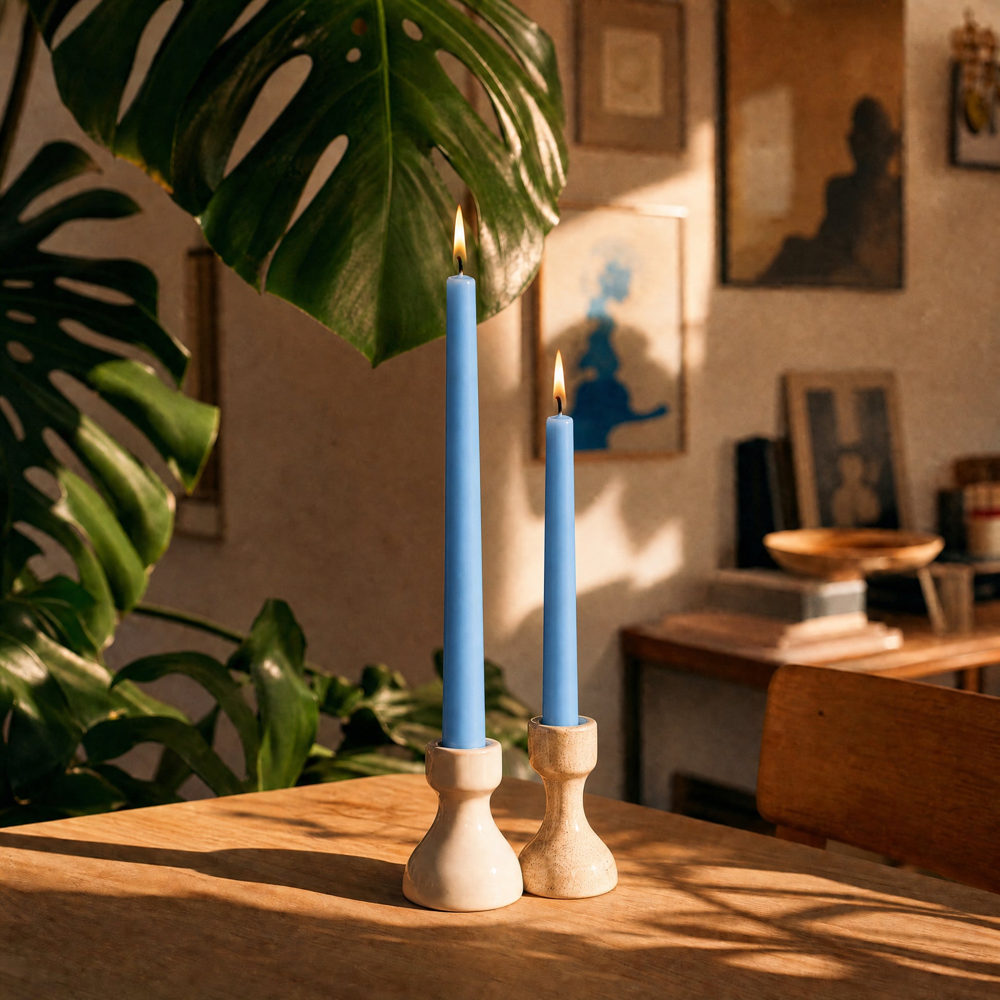 Two blue beeswax taper candles in wooden holders on a table with plants and a warm interior setting.