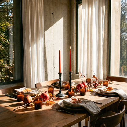 Dining table set for a meal with dark orange beeswax taper candles and autumn decorations in a bright room with large windows.