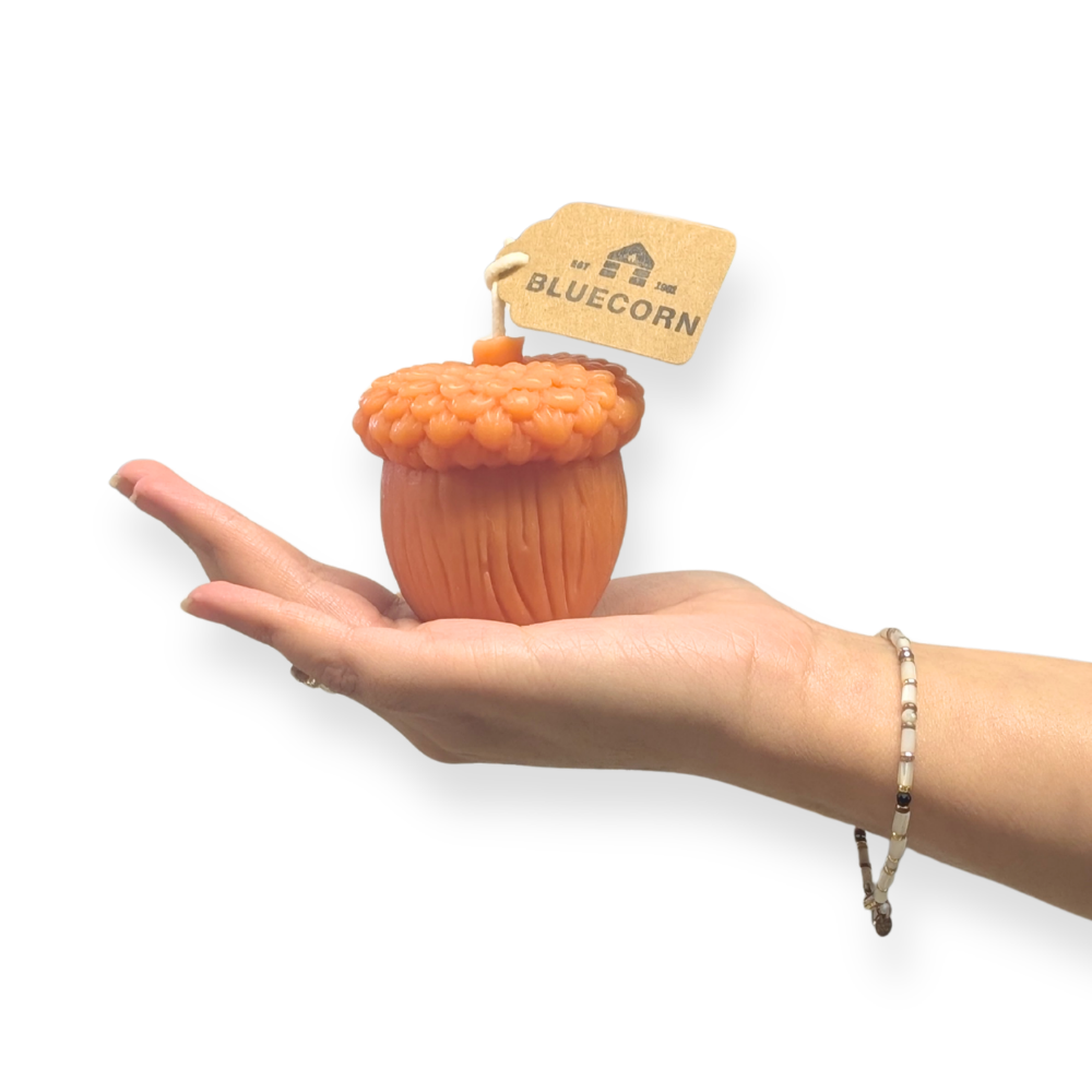 Raw Acorn shaped candle being held in a hang to show size, with Bluecorn tag and white background